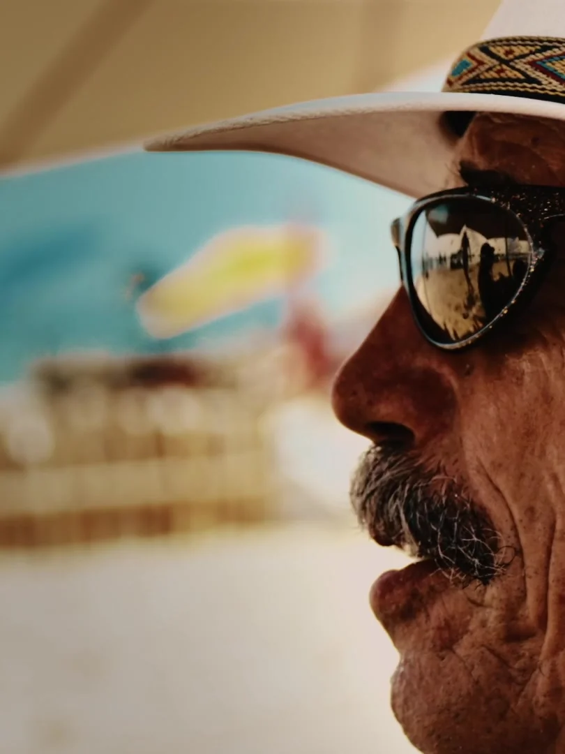 Perfil cercano de un hombre mayor con sombrero y gafas de sol que reflejan la playa.
