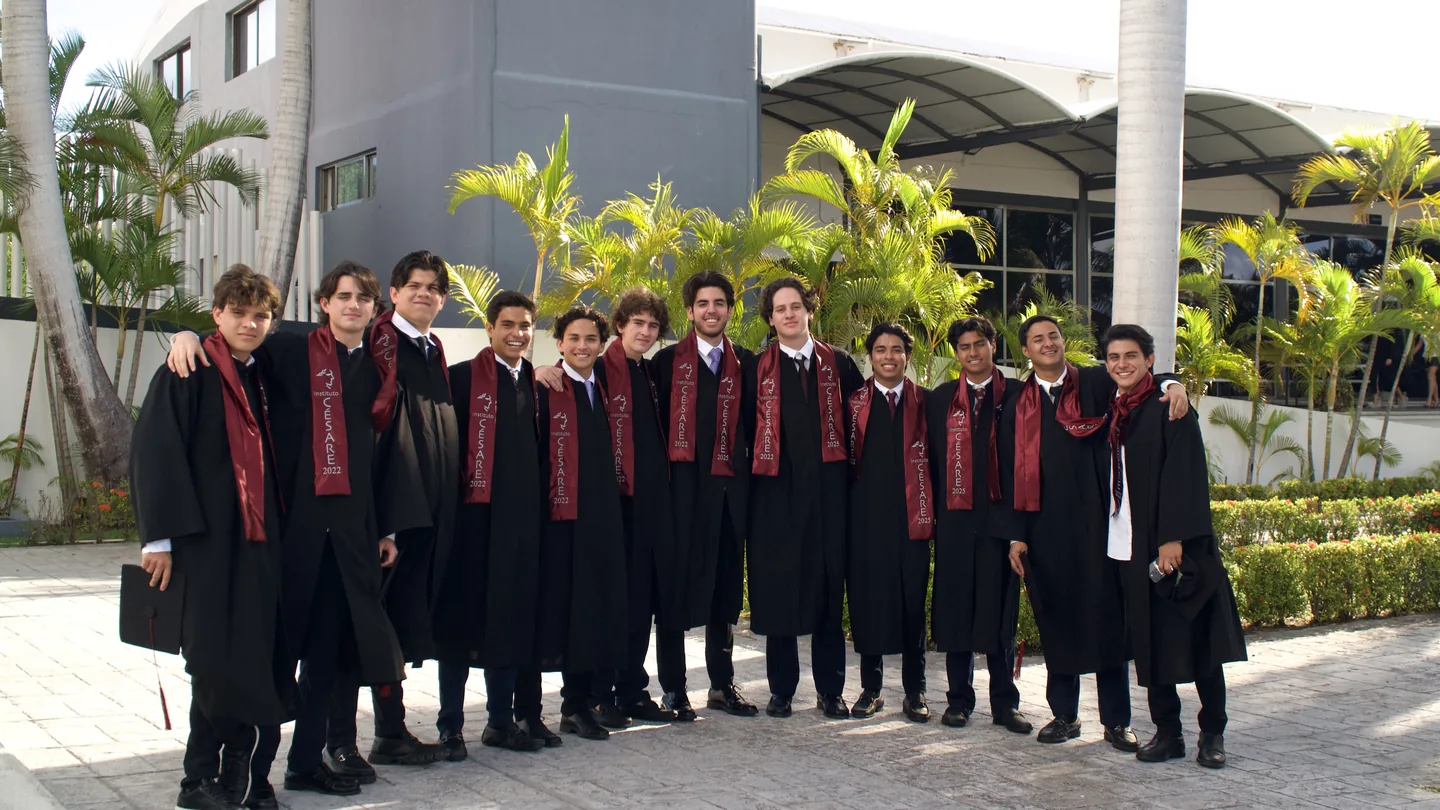 Grupo de graduados con togas negras y bandas rojas frente al edificio escolar.