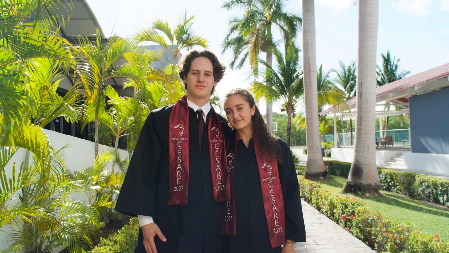 Pareja de graduados con bandas rojas entre palmeras frente al campus.