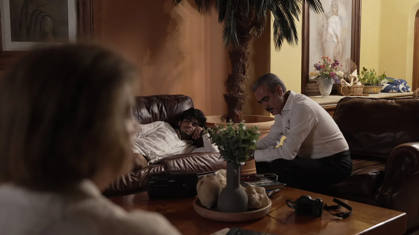 Escena íntima en la sala: figura mayor junto a alguien recostado en el sofá.