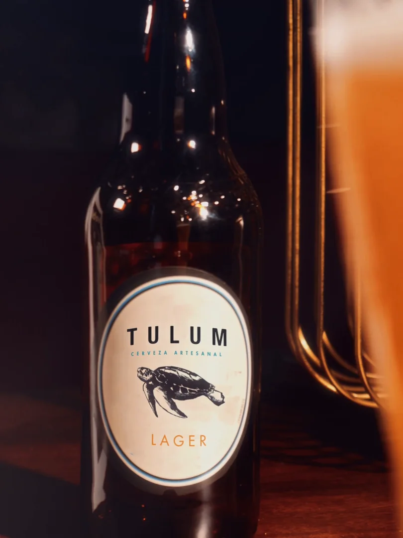 Botella de Tulum Lager a foco, con vaso borroso a la derecha y el borde de una foto enmarcada.