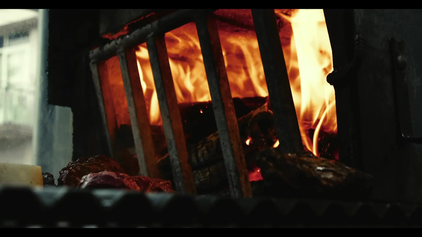 Brasas y llamas dentro de un asador de hierro con carne en primer plano desenfocado.