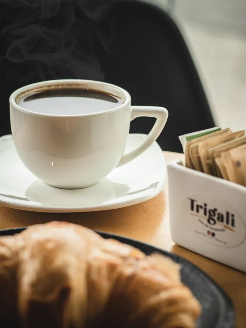Taza blanca humeante junto a un croissant y servilleta Trigali.